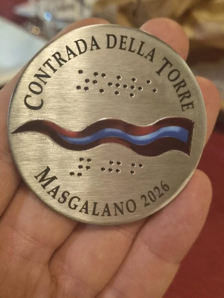 medaglia metallica celebrativa circolare: Riporta le scritte incise "CONTRADA DELLA TORRE" lungo il bordo superiore e "MASGALANO 2026" lungo quello inferiore. Al centro è presente un'onda con i colori della Contrada della Torre (rosso bordeaux, blu e bianco). Sopra e sotto il motivo colorato sono incisi i punti dell'alfabeto Braille, CON SCRITTO TORRE 2026