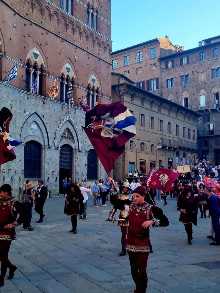 Corteo Storico in Piazza del Campo: Gli alfieri e i figuranti della Contrada della Torre in abiti storici rinascimentali mentre sventolano le bandiere nei colori bordeaux, bianco e blu davanti al Palazzo Pubblico.