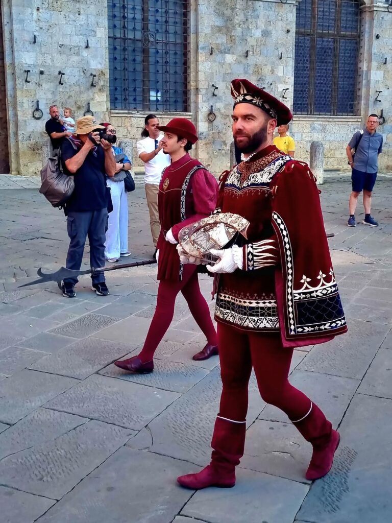 Trasporto del Masgalano: Un figurante della Contrada della Torre in abito di velluto finemente ricamato che trasporta con cura la scultura del Masgalano durante la sfilata.