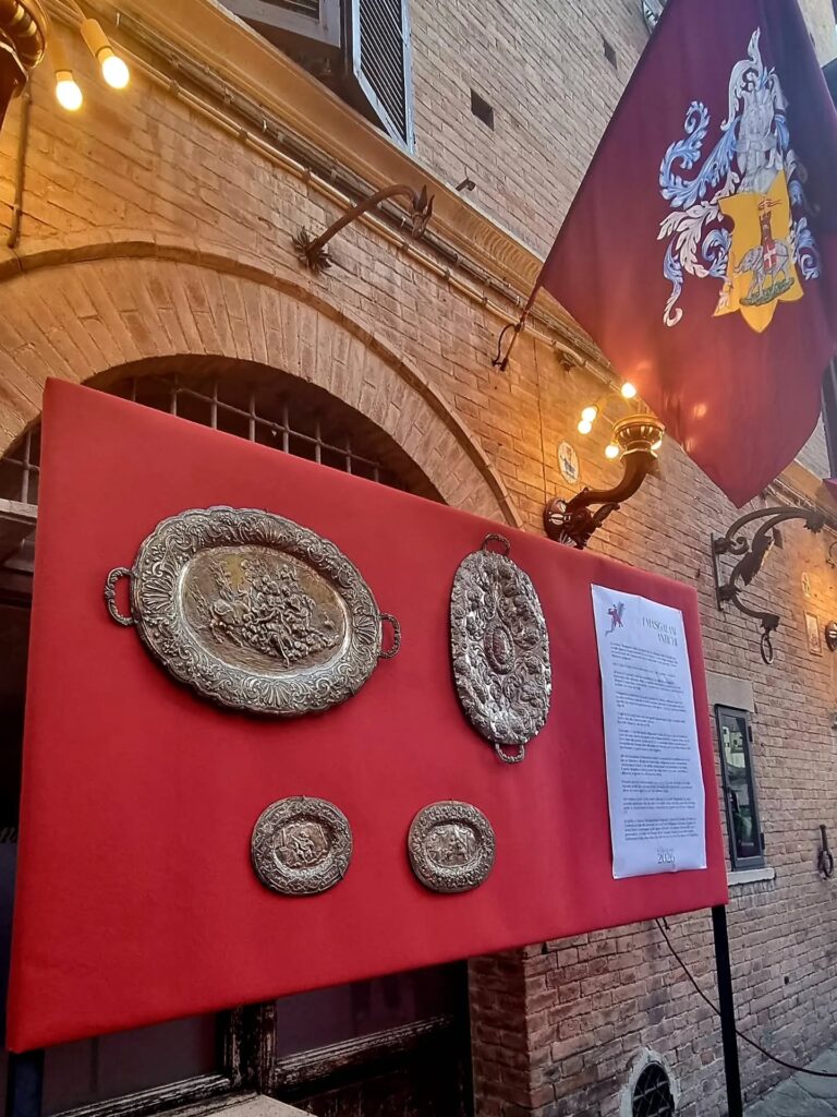 Esposizione dei MASGALANI storici : Un tabellone allestito all'esterno rivestito di panno rosso su cui sono esposti antichi MASGALANI in argento, affiancato dalla bandiera con lo stemma della Contrada della Torre.