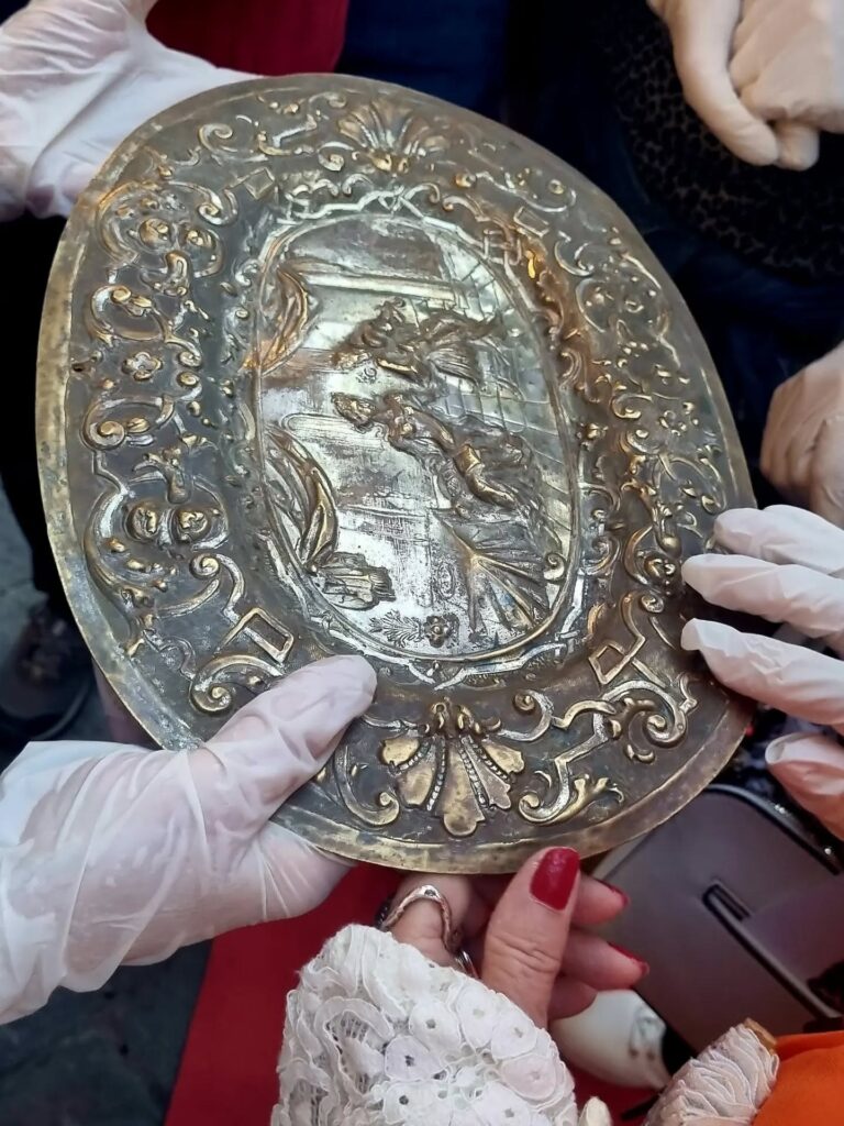 Dettaglio del MASGALANO antico: Mani protette da guanti bianchi che sostengono un antico MASGALANO A FORMA DI PIATTO OVALE in argento cesellato a sbalzo con raffigurazioni figurative.