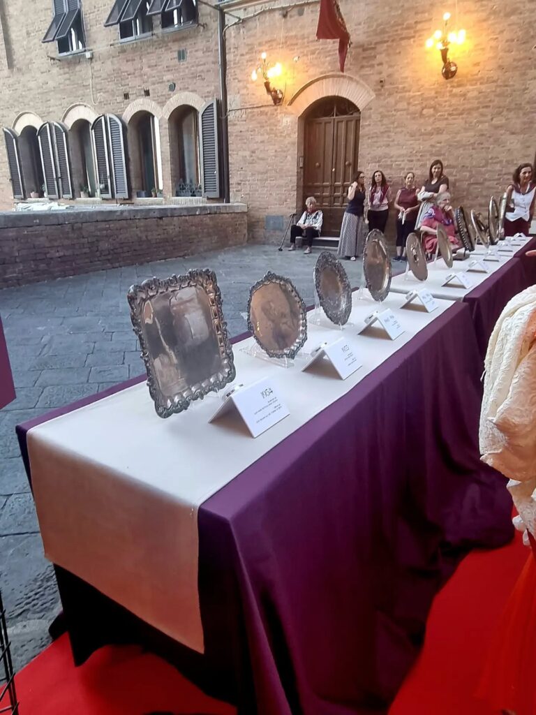 Mostra dei Masgalani del passato : Una lunga tavola allestita all'aperto con drappo viola su cui sono esposti in sequenza cronologica i vari MASGALANI d'argento vinti nel tempo, con appositi cartellini esplicativi.
